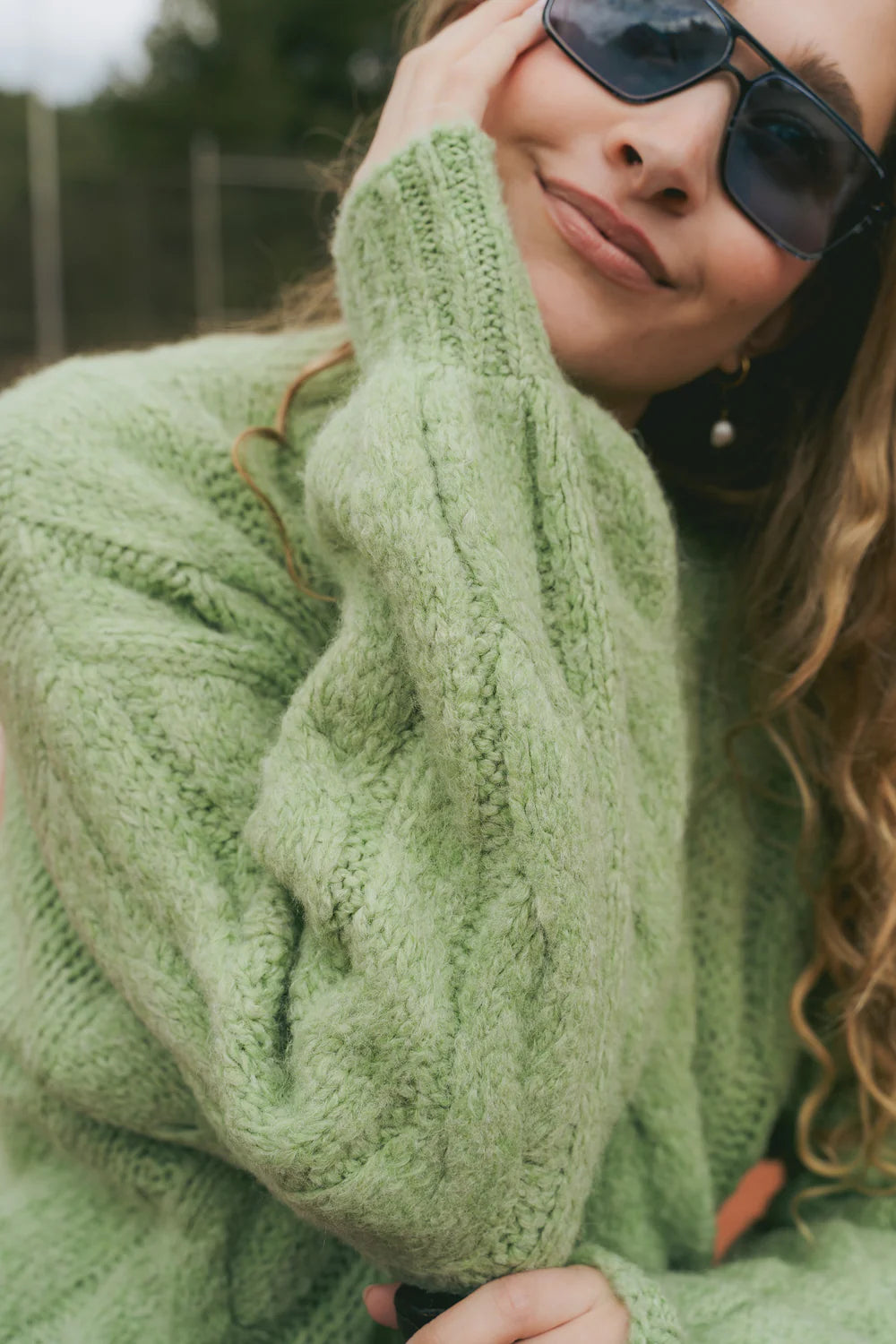 Person wearing a MindTraveler Chiara green knitted sweater and sunglasses with a blurred background