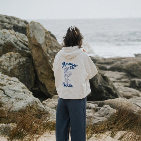 Person wearing a hoodie with text and graphics, standing on a rocky beach.