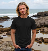 Man wearing a black t-shirt standing on a rocky beach with ocean in the background